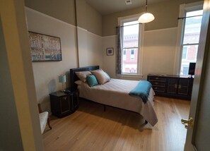 A bedroom with a bed, bedside table, lamp, and a window with a view of buildings.