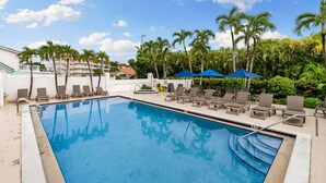 A swimming pool with lounge chairs and umbrellas, surrounded by palm trees and a clear sky.