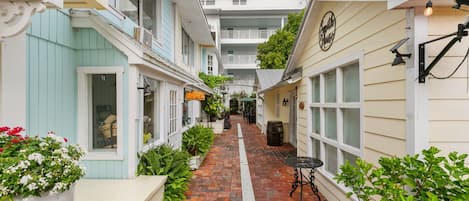 Un étroit sentier pavé de briques serpente entre les immeubles, qui sont dotés de balcons, et on y trouve une petite table accompagnée d’une chaise.