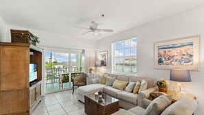 A spacious living room with a large sofa, a coffee table, and a television. There is a balcony with a view of the city.