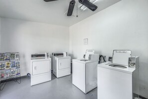 Une buanderie équipée de quatre machines à laver et de quatre sèche-linge blancs, d’un ventilateur de plafond et d’un tableau d’affichage où sont accrochées diverses annonces.