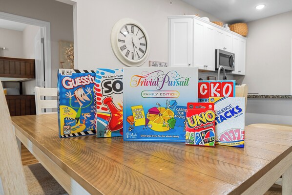 Uma mesa de madeira com vários jogos de tabuleiro, incluindo Advinhando Quem, So So, Trivia Pursuit, UNO e Sequence Tactics, em um quarto com um relógio e armários.