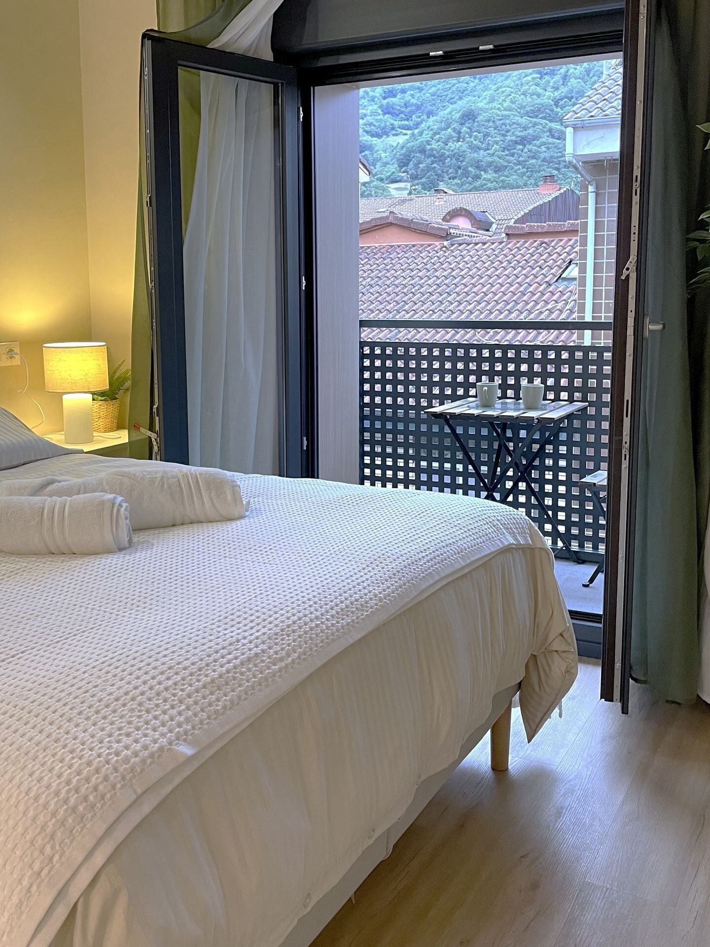 A bedroom with a bed, a nightstand, a lamp, and a view of a balcony with a table and chairs.