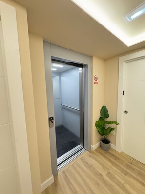 An elevator with a handrail, a no-smoking sign, and a potted plant beside a door. - SJ Puerta de Redes (Pola de Laviana)