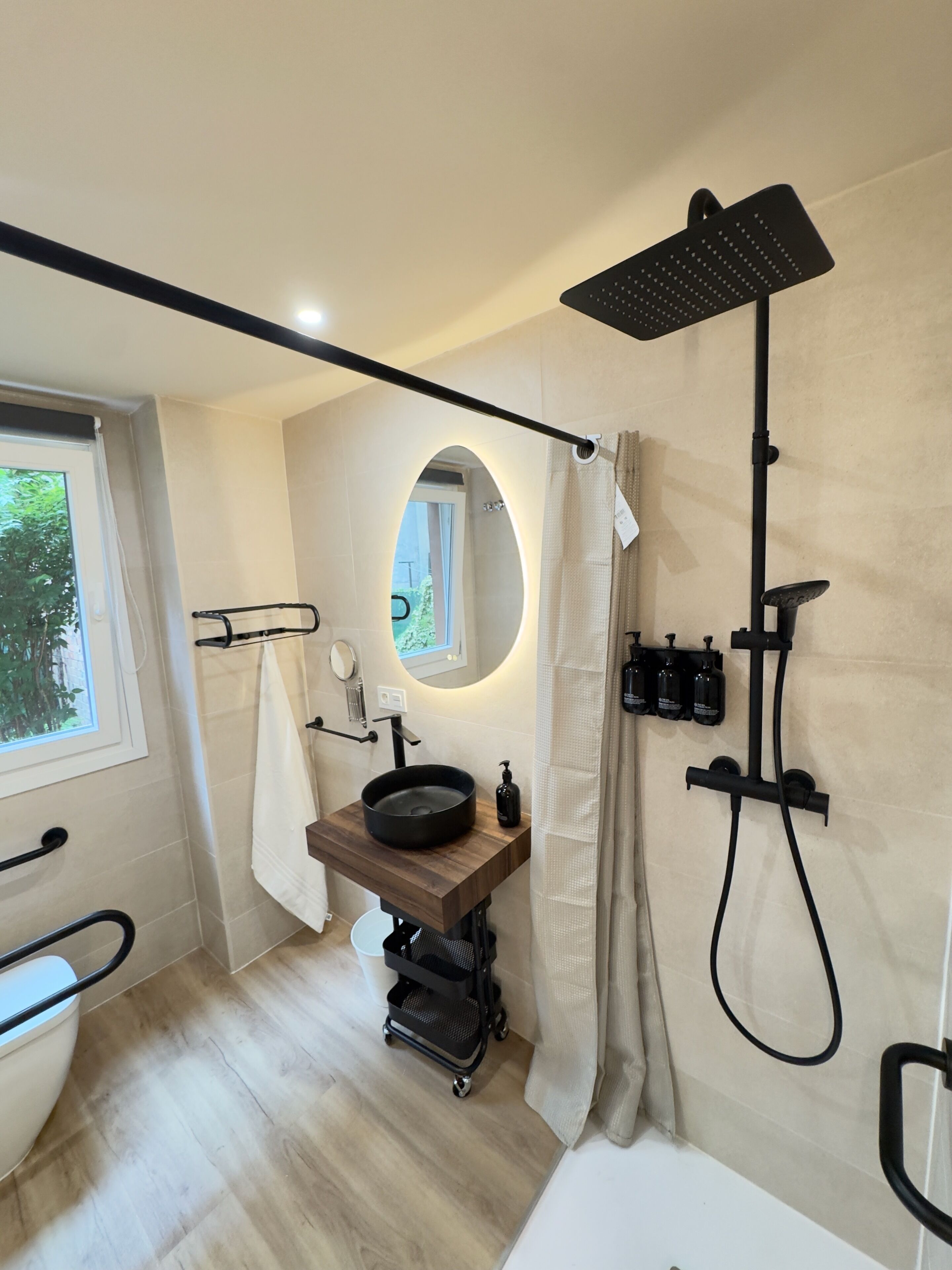 A modern bathroom with a wooden vanity, a round mirror, a black sink, a handheld showerhead, and a toilet.