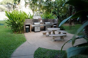Eine Terrasse im Freien mit Grill, Steintisch und einer Sitzgelegenheit, umgeben von Grünpflanzen.