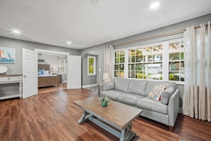 Ein geräumiges Wohnzimmer mit grauem Sofa, Holztisch und einem großen Fenster mit Vorhängen.