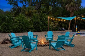 A fire pit surrounded by four Adirondack chairs, with string lights overhead and a backdrop of dense foliage. - Heated Pool + Spa, Fenced Yard, Fire Pit, Sleeps 8, 15 Min to AMI (Bradenton)