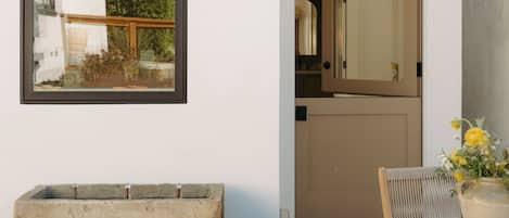 A room with a concrete bench, a wooden chair, a table with a vase, a framed picture, and a door leading to a kitchen.