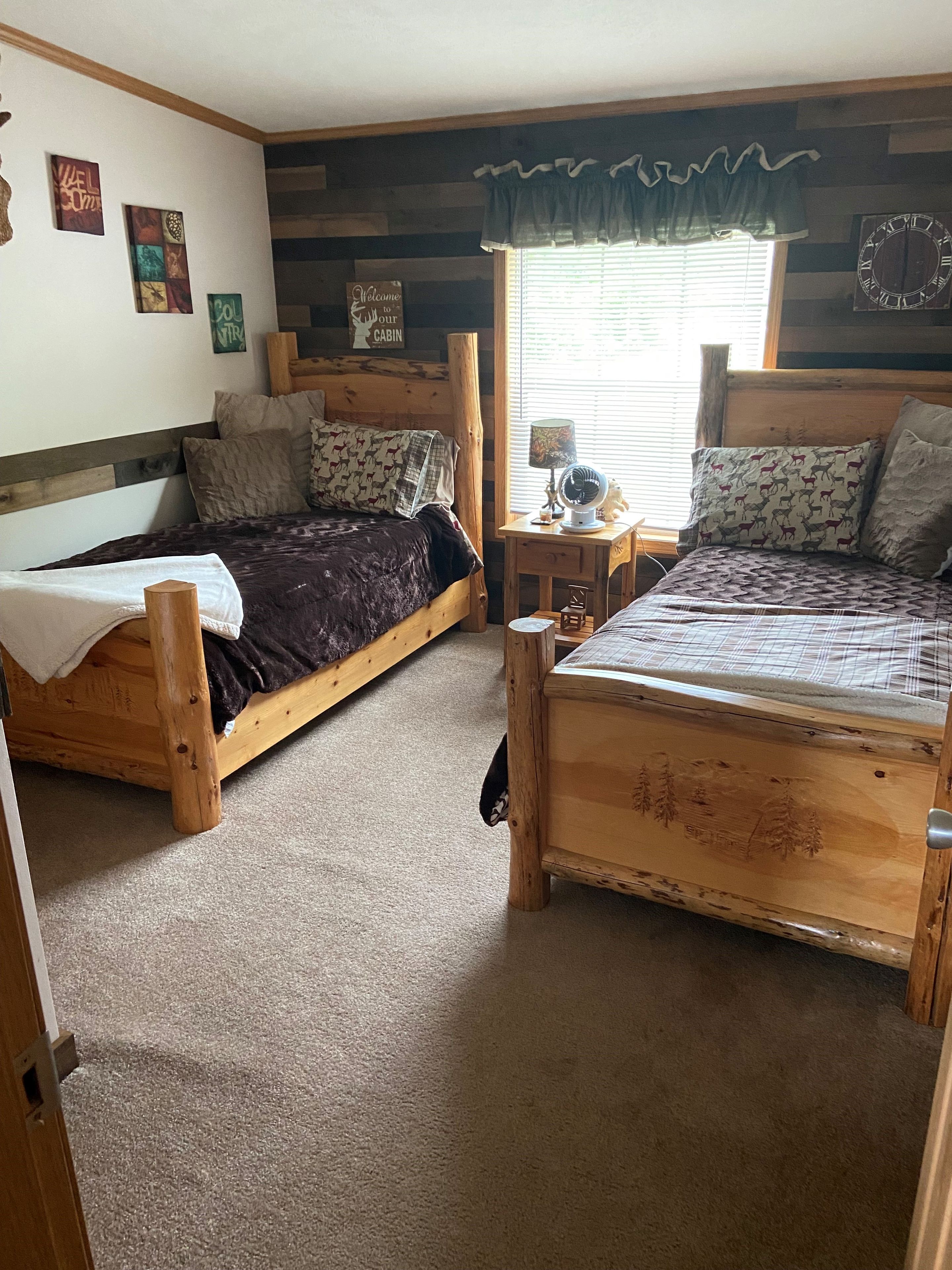 Une chambre de style cabane avec deux lits en bois, une petite table avec une lampe et une fenêtre avec des stores.