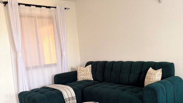 Ein Wohnzimmer mit einem dunkelgrünen Sofa, einem kleinen runden Tisch, einem Deckenventilator und einem Fenster mit durchsichtigen Vorhängen.