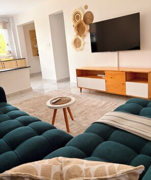A modern living room with teal sofas, a small round table, a flat-screen TV mounted on the wall, and a wooden cabinet below. - Modern 1-Bedroom Coastal Apartment in Mtwapa Near the Beach. (Mtwapa)
