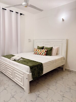 A white bed with a green throw blanket, white curtains, and a ceiling fan. - Modern 1-Bedroom Coastal Apartment in Mtwapa Near the Beach. (Mtwapa)