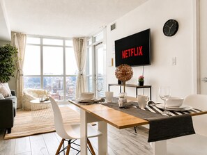 A modern living room with a dining area, a large window with curtains, a flat-screen TV displaying 'NETFLIX', and a city view.