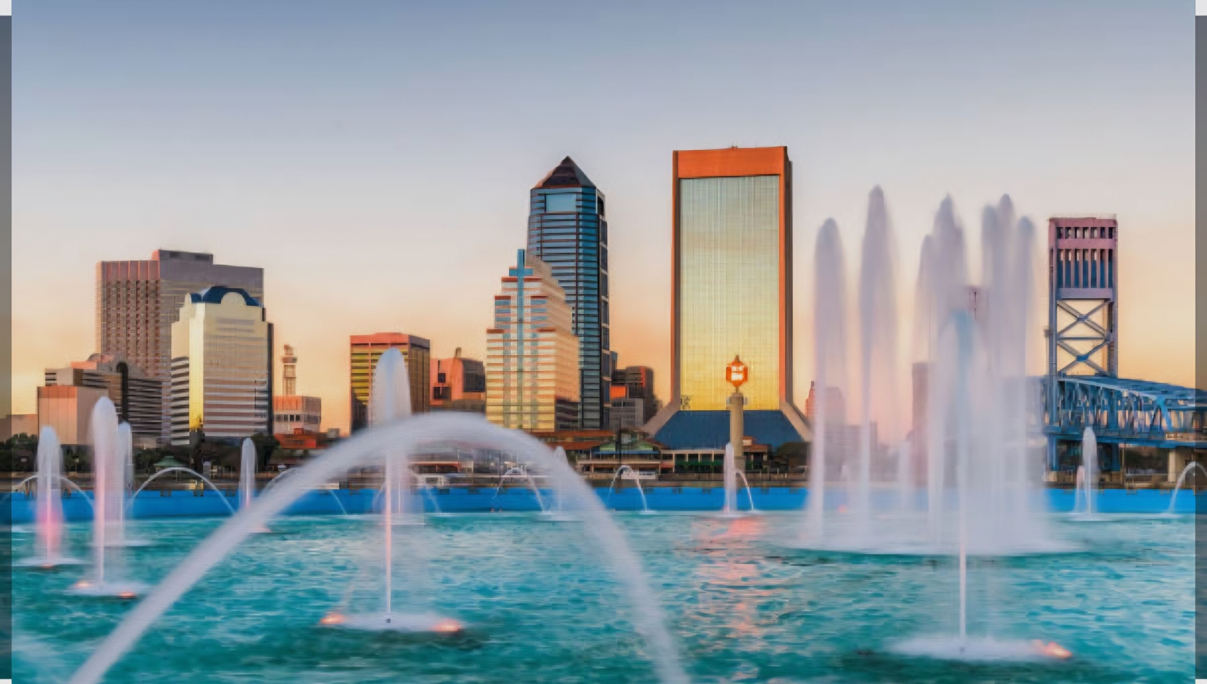 A city skyline with modern buildings and a bridge, and water fountains in the foreground.
