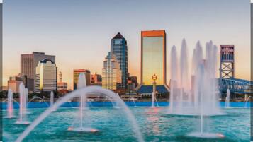 A city skyline with modern buildings and a bridge, and water fountains in the foreground.