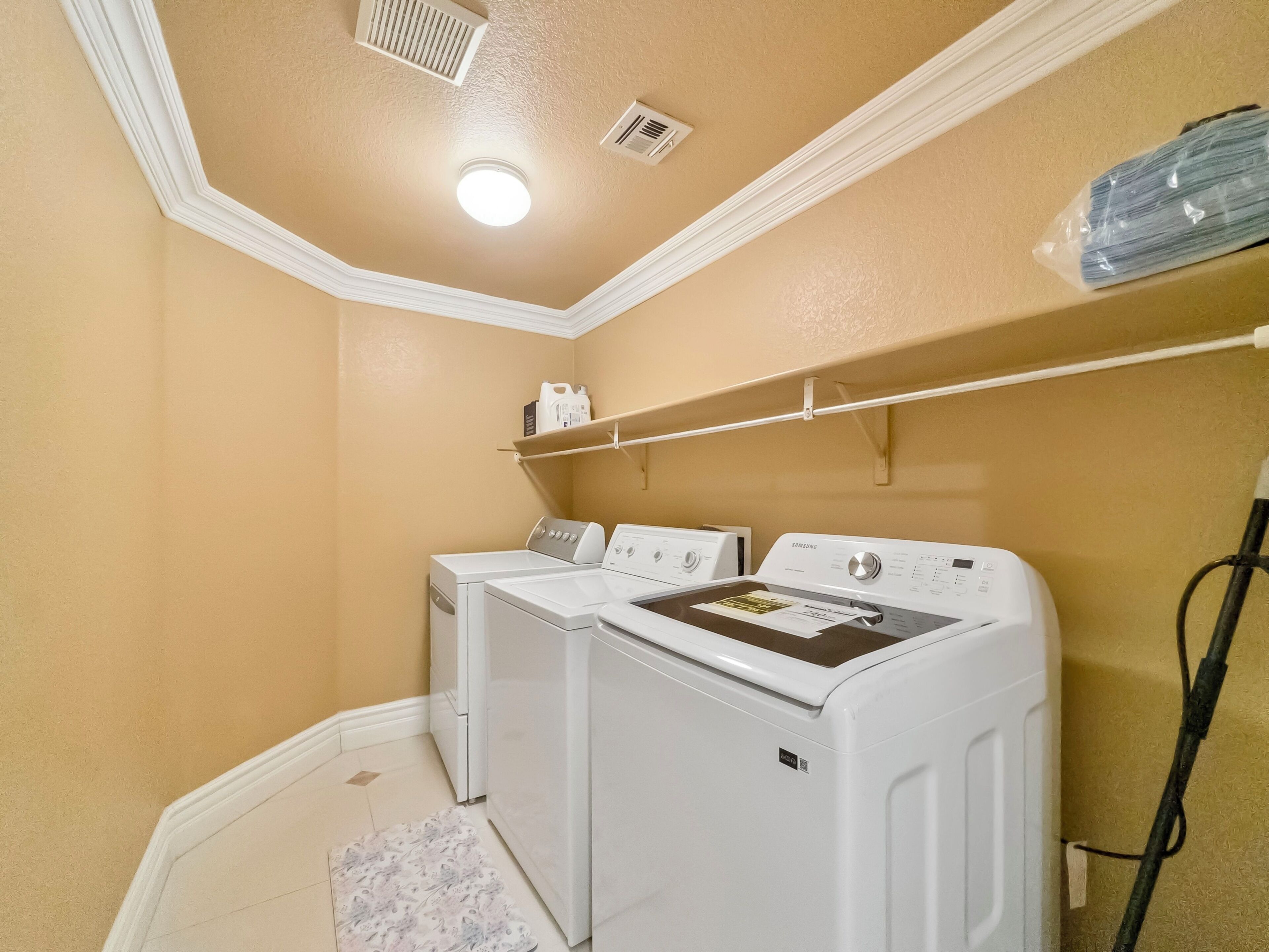 A laundry room with two white front-loading washers and dryers, a ceiling vent, a shelf with laundry supplies, and a power cord.