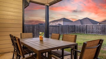 Un patio techado con una mesa y sillas de madera, un florero con flores y vistas a una valla de madera y casas al fondo.