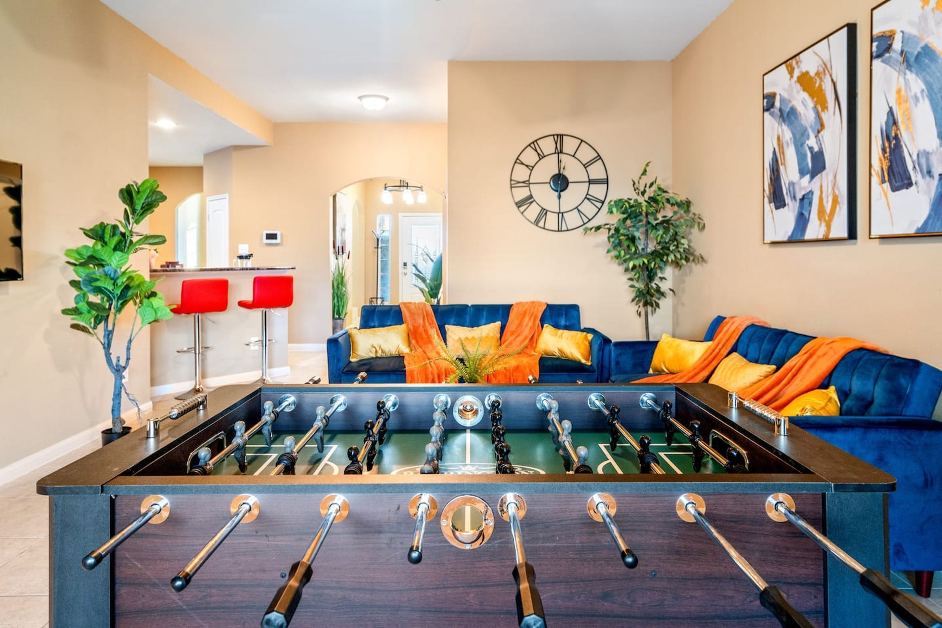 Une pièce avec une table de baby-foot, un canapé bleu avec des coussins orange, une horloge au mur et un miroir à l’arrière-plan.