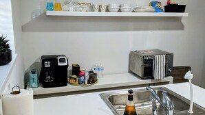 Una cocina moderna con encimeras blancas, un lavamanos de acero inoxidable, una cafetera, un tostador y una pequeña planta en maceta.