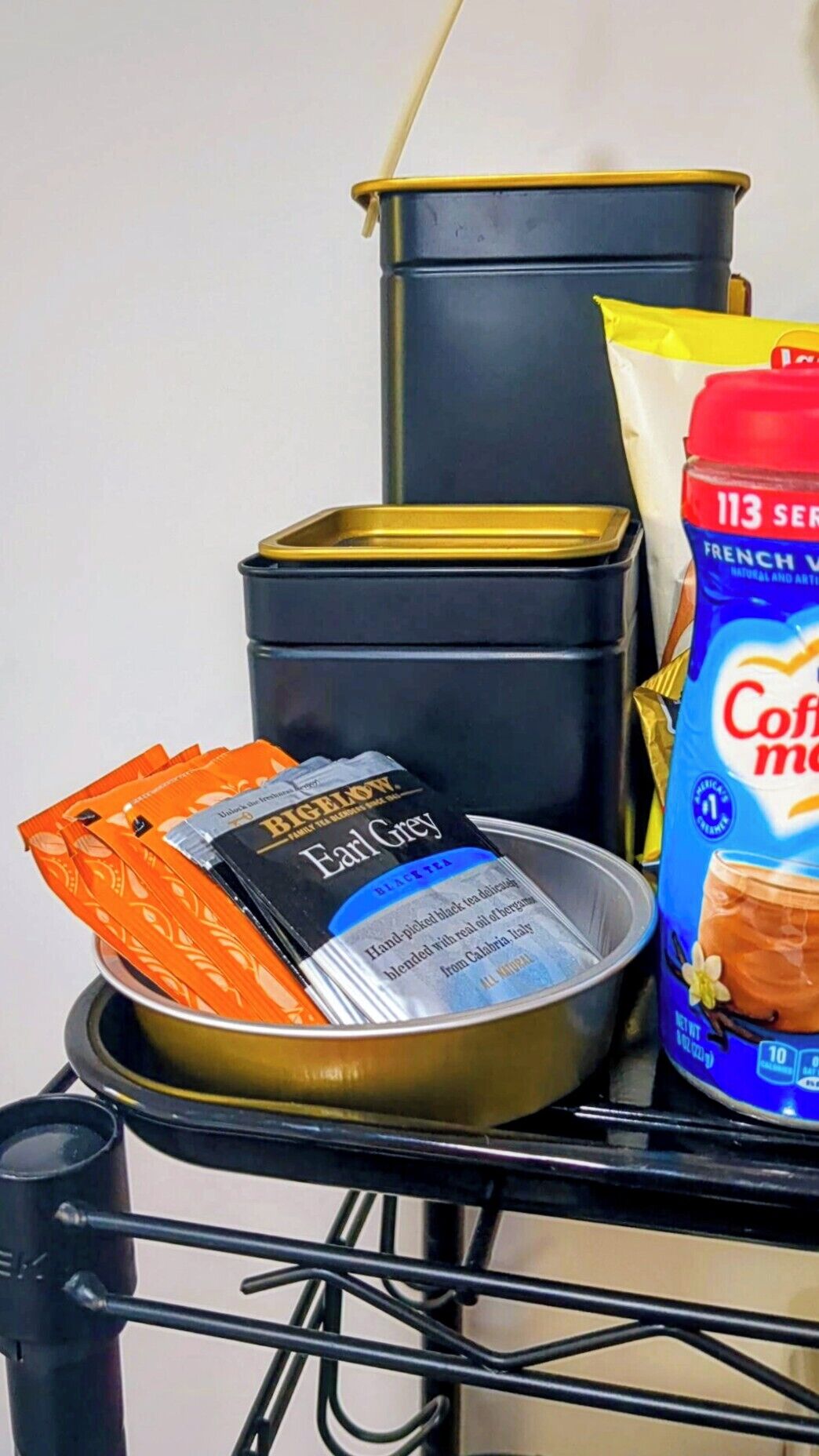 A shelf with a black container, a tin of Earl Grey tea, a bag of coffee mix, and a can of French vanilla creamer.