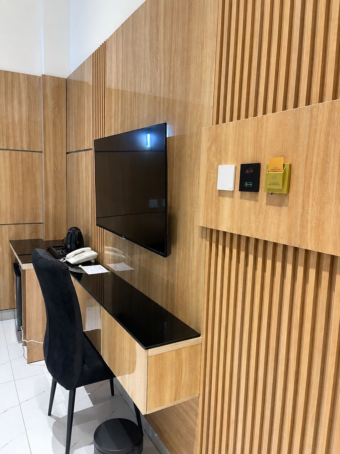 Una habitación de hotel moderna con pared de paneles de madera, un televisor de pantalla plana sobre un escritorio, una silla negra y una mesita con un teléfono y papeles.