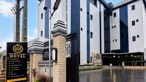 Un hotel moderno con una entrada destacada, un cartel que anuncia servicios como restaurante, piscina y gimnasio, y un área pavimentada bien cuidada al frente.