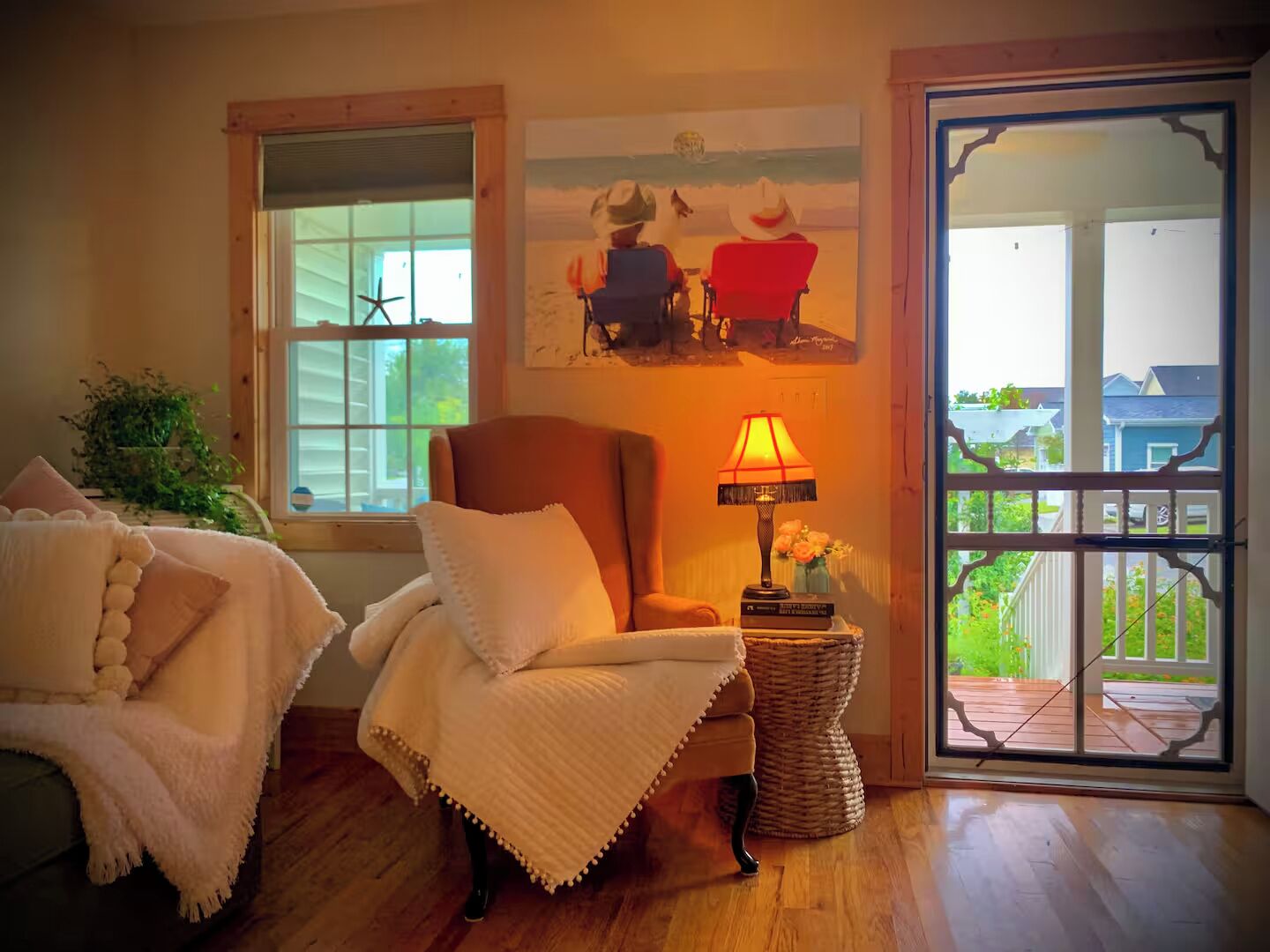 A cozy room with a wooden floor, a comfortable armchair, a small table with a lamp, and a view of the outdoors through a glass door.