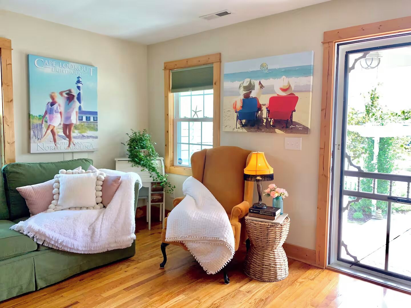 Una habitación con un sofá verde, un sillón blanco, una mesita con una lámpara y obras de arte que representan escenas de playa.