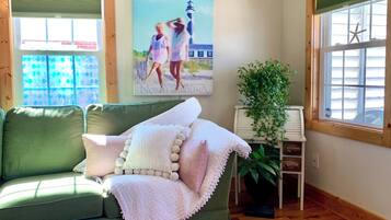 A room with a green sofa, a white blanket, a framed picture of a lighthouse, a potted plant, and a window with blinds.