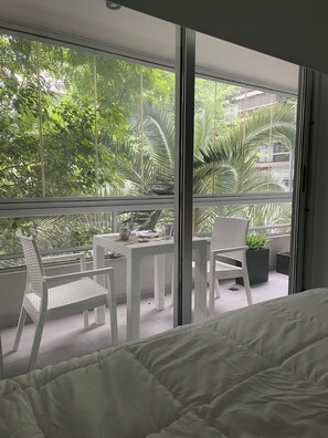 A balcony with a white table and chairs, a bed, and a view of greenery outside. - Premium Arcos Apartment. Modern. In the safest area of Buenos Aires. (Buenos Aires)