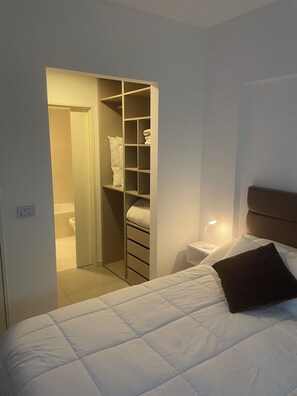 Ein Schlafzimmer mit Bett, Nachttisch, Nachttischlampe, Kopfteil und begehbarer Kleiderschrank mit Regalen.