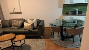 A living room with a sofa, two round wooden tables, a glass-top dining table with chairs, a kitchen with white cabinets and a green backsplash, and a patterned rug. - Superb 2-bedroom apartment in adorable Windhoek neighborhood (Windhoek)