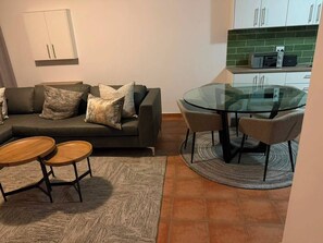 Ein Wohnzimmer mit einer Couch, zwei runden Holztischen, einem Esstisch mit Glasplatte und Stühlen, einer Küche mit weißen Schränken und einer grünen Spritzschutzwand sowie einem gemusterten Teppich.