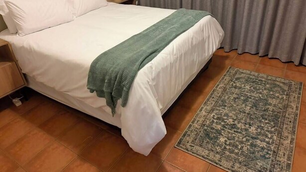 A neatly made bed with white linens and a green throw blanket, a bedside table with a lamp, a patterned rug on the floor, and a grey curtain in the background. - Superb 2-bedroom apartment in adorable Windhoek neighborhood (Windhoek)