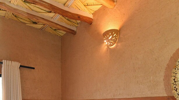 Ein Einzelbett mit weißem Kopfkissen und roter Decke, eine Holzbalkendecke, eine an der Wand befestigte Leuchte und ein Vorhang.