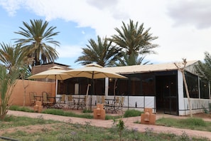 An outdoor dining area with tables and chairs, palm trees, and a building with large windows. - Ferme Dar Charach (Tarmigt)