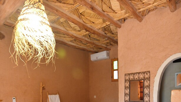 A room with a thatched ceiling, a bed, a small table, a ladder, and a wall-mounted air conditioner. - Ferme Dar Charach (Tarmigt)