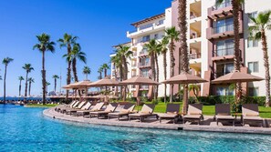 Ein Hotelpool mit Liegestühlen und Sonnenschirmen, umgeben von Palmen und einem mehrstöckigen Gebäude.