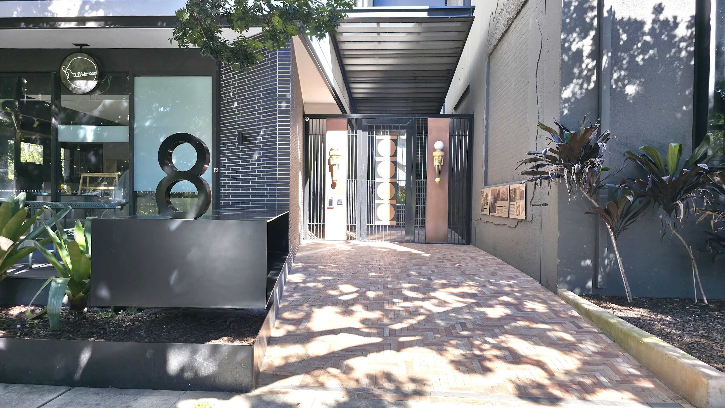 A modern building entrance with a brick pathway, metal gates, and outdoor seating areas.