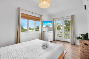 A hotel room with a bed, a television, a dresser, and a balcony with a view of palm trees. - Sea Haven | A Charming Florida Keys Cottage Near Sombrero Beach (Marathon)