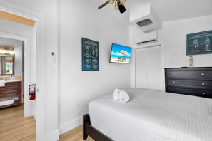 Ein Schlafzimmer mit einem Bett, einem Schminktisch, einem Deckenventilator, einem an der Wand montierten Fernseher und einem Bild von einer Strandlandschaft.