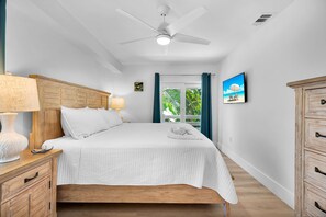 A bedroom with a bed, a ceiling fan, a nightstand with a lamp, a dresser, and a wall-mounted TV displaying a beach scene. - Sea Haven | A Charming Florida Keys Cottage Near Sombrero Beach (Marathon)