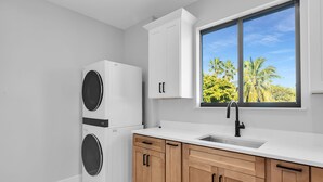 A kitchen with a built-in washer and dryer, a window with a view of palm trees, and wooden cabinets. - Coco Plum Palms New Waterfront Pool Spa Dock (Marathon)