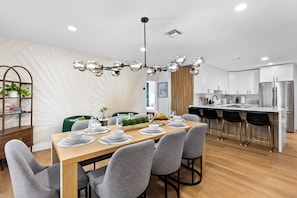 A modern kitchen with white cabinets, a stainless steel refrigerator, and a dining area with a wooden table set for four. - Quiet Oasis-Heated Pool •Fire Pit •Outdoor Games (Fort Lauderdale)