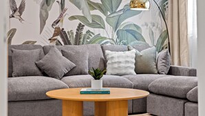 Ein Wohnzimmer mit grauem Sofa, einem runden Holztisch, einer Topfpflanze und einem Buch mit dem Titel 'COASTLINES'.