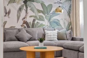 Ein Wohnzimmer mit grauem Sofa, einem runden Holztisch, einer Topfpflanze und einem Buch mit dem Titel 'COASTLINES'.