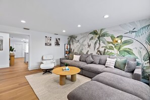 A modern living room with a grey sectional sofa, a white armchair, a round coffee table, and a large wall mural of tropical foliage. - Quiet Oasis-Heated Pool •Fire Pit •Outdoor Games (Fort Lauderdale)