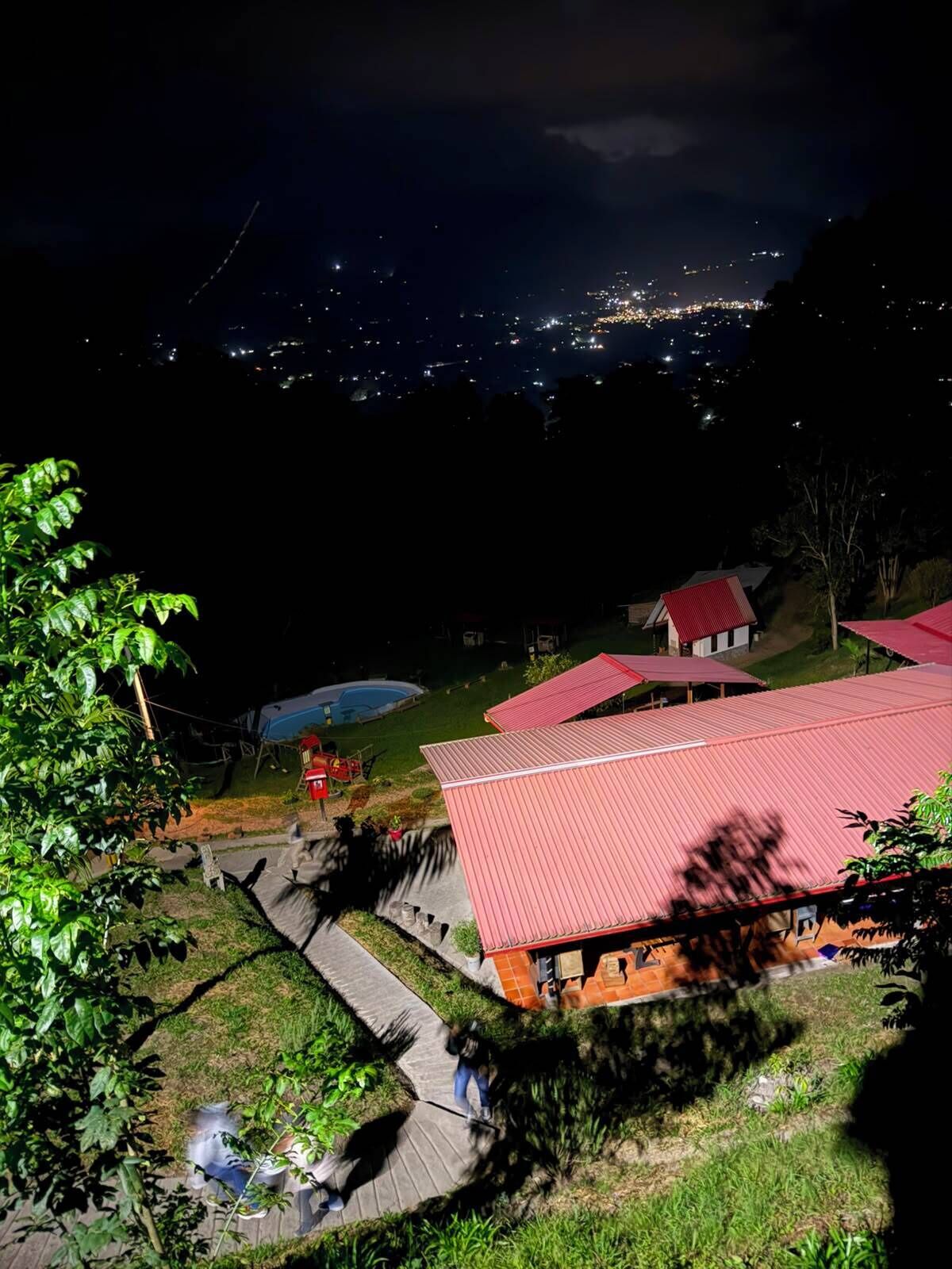 A nighttime view of a hillside with a red-roofed building, a swimming pool, and a paved walkway.
