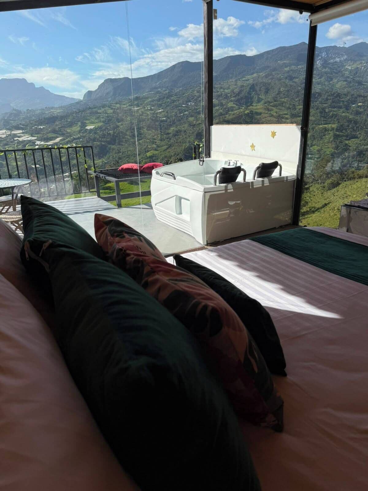 Un dormitorio con una cama grande, vistas a montañas, un balcón con mesa y sillas, y un jacuzzi.